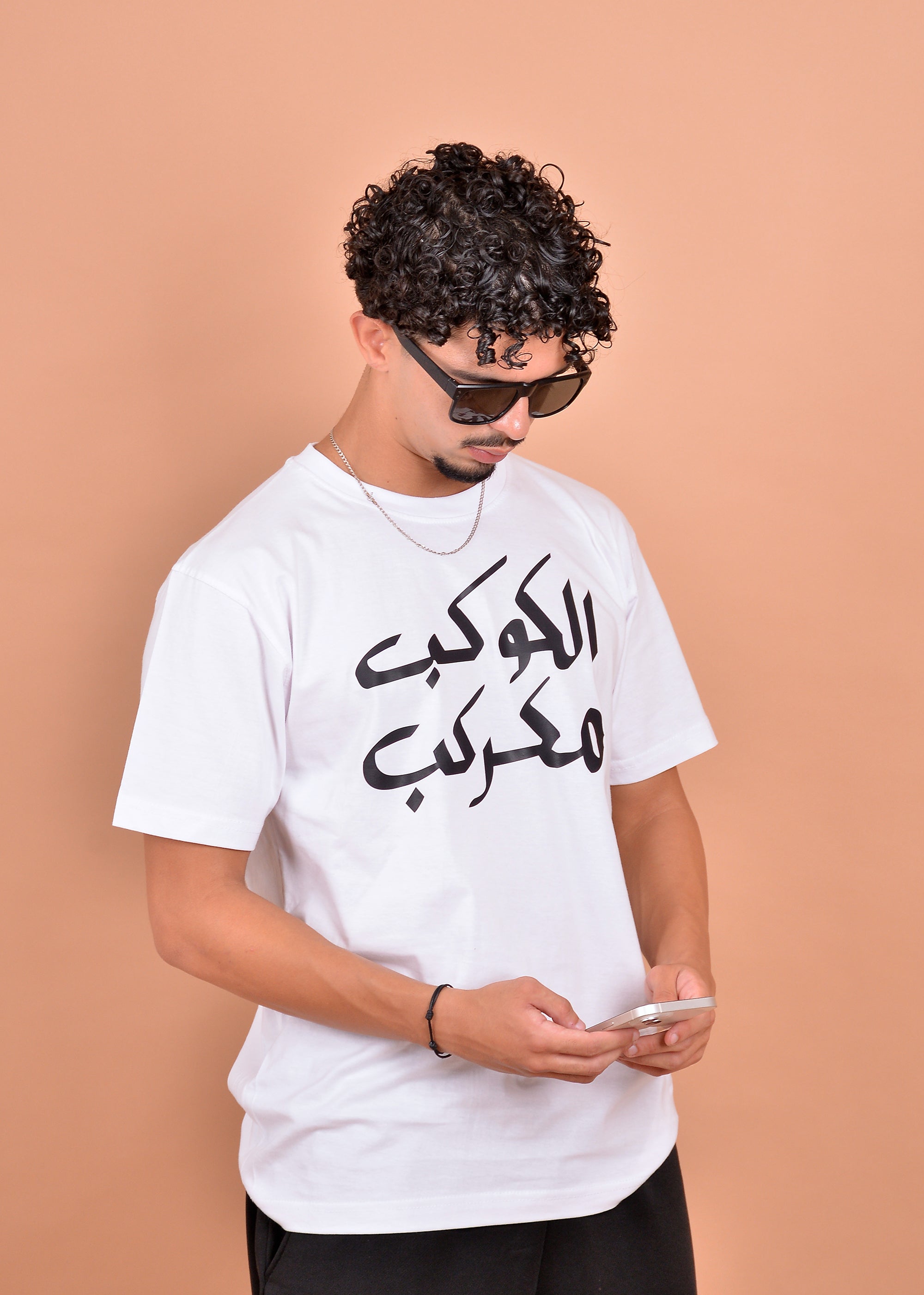 Man wearing a white t-shirt with Arabic text, using a phone on a beige background